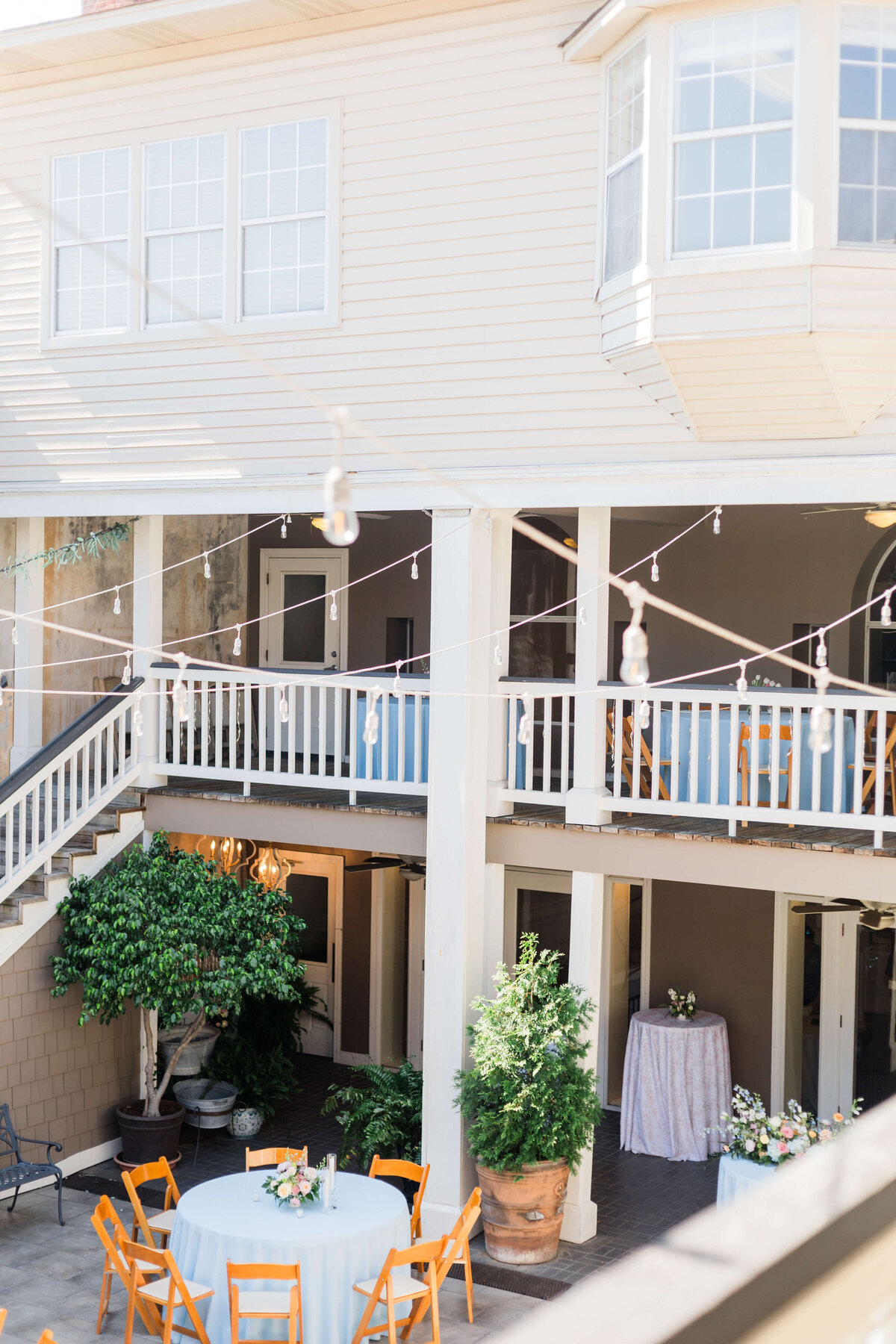 A courtyard with round tables covered in light blue tablecloths, wooden chairs, potted plants, and string lights, surrounded by a two-story beige balconies with white railings and staircases.