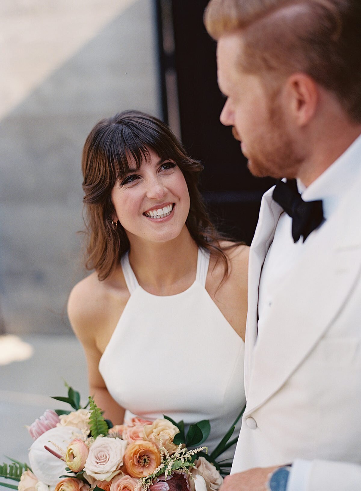 The bride, a brunette wearing a halter top sheath wedding dress, holding a large bouquet of peach, blush and pink flowers smiles up at the groom who is wearing a tuxedo with a white jacket and bow tie.