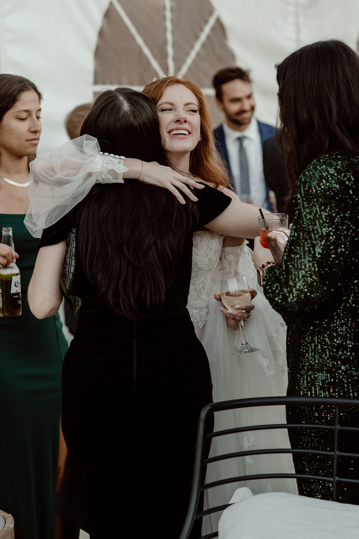 Bride joyfully hugs a guest at her wedding reception, surrounded by friends and family. The bride, holding a glass of wine, wears a lace wedding gown, while guests in elegant attire celebrate the occasion.