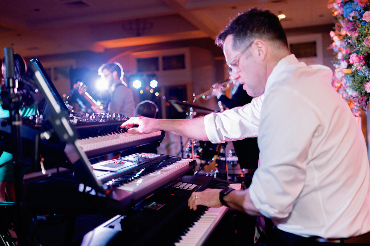 A musician plays the keyboard during Callie & Hayes’ wedding reception at Burritt on the Mountain, with vibrant stage lights and other band members performing in the background.