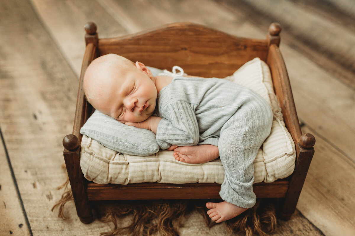 Baby boy in a light blue PJ outfit laying on a tiny wooden baby bed.