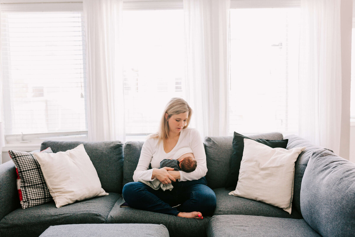 Mom sits on the couch to nurse her newborn baby in a light filled room