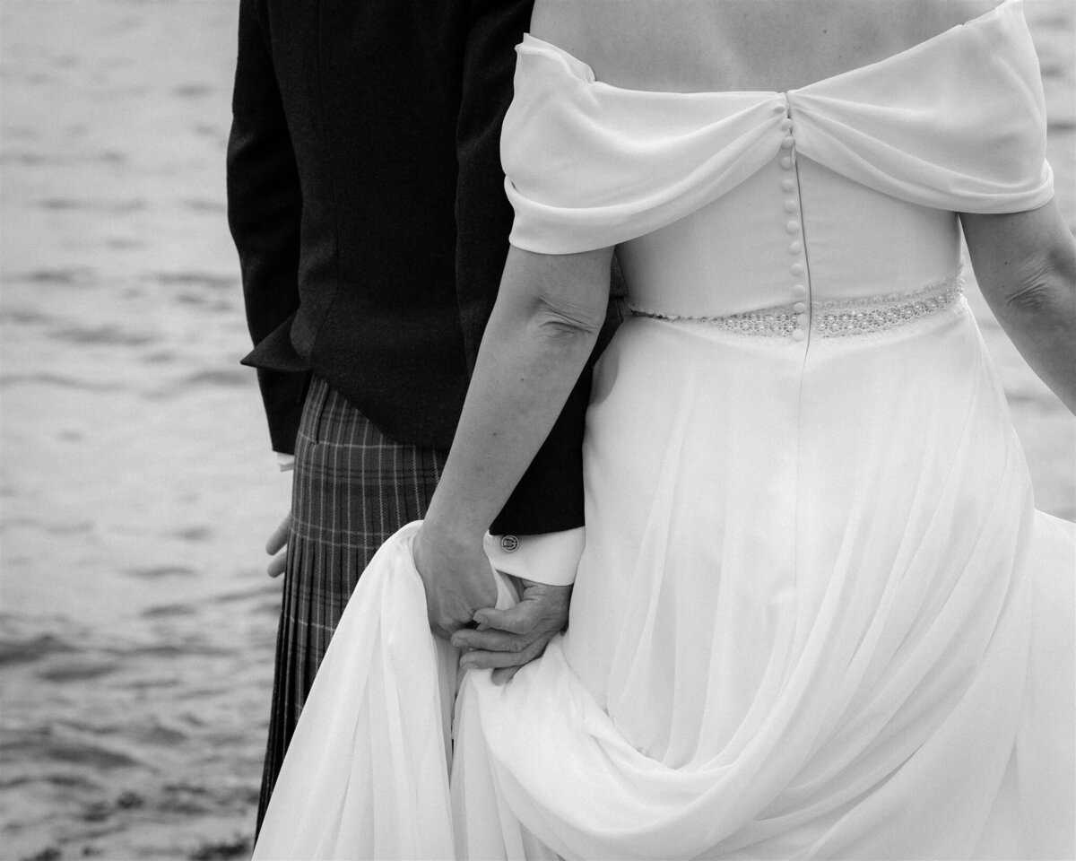 close up a bride and groom holding hands behind their back as they stand by the sea