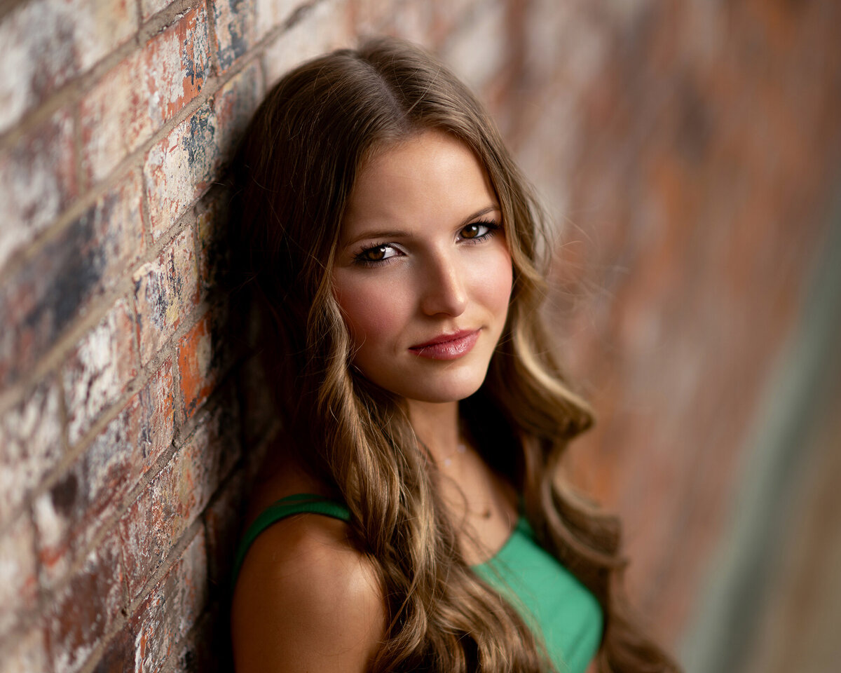 A brunette in a green dress leans on a brick wall smiling after attending private schools in Iowa