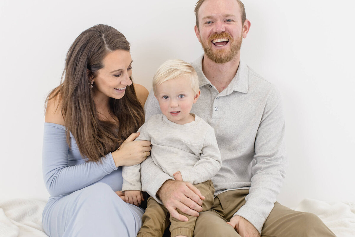 A smiling family sits close together against a white background. The woman, dressed in a light blue off-shoulder dress, sits beside the man in a gray shirt and khaki pants. A small child with blond hair sits on the mans lap, wearing a beige outfit.