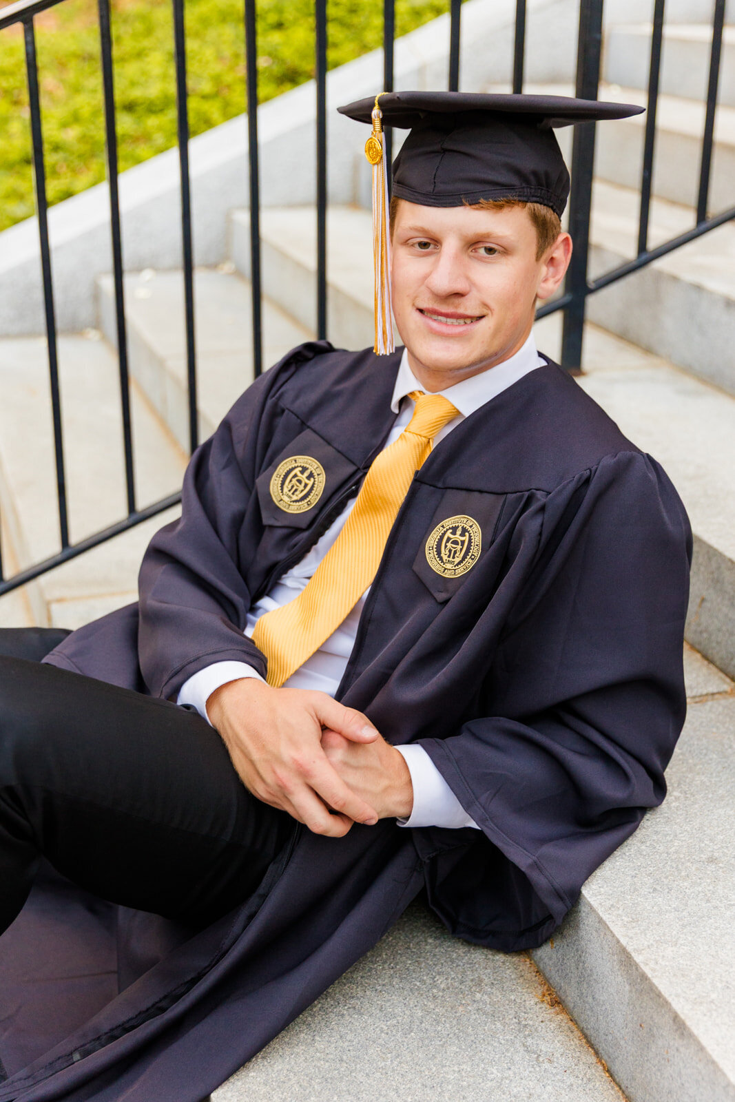 GT senior boy sitting on the campus tower stairs leaning back and wearing graduation outfits cap and gown with a yellow tie during senior portraits with Atlanta photographer Laure Photography