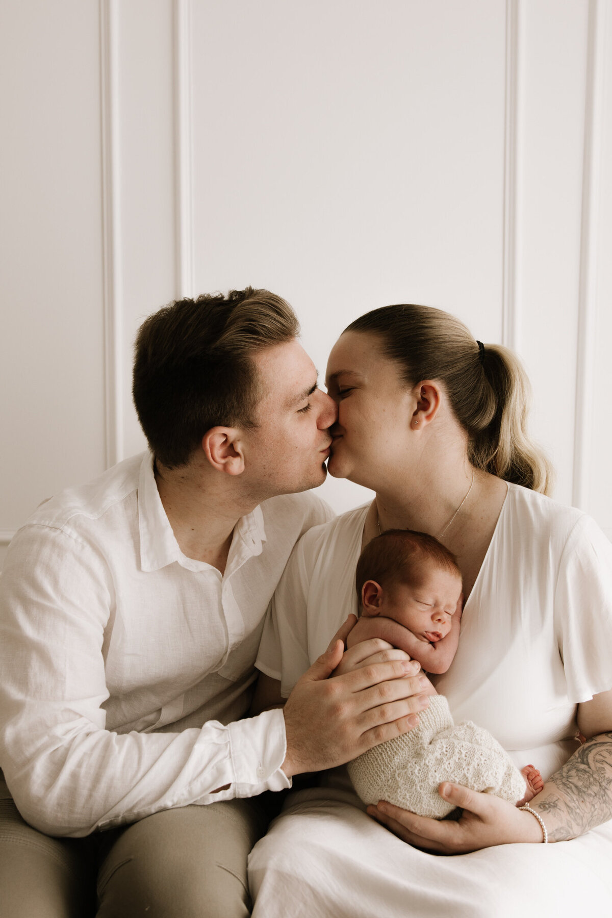 A tender moment during a newborn session where two parents, dressed in soft neutrals, share a gentle kiss while holding their sleeping baby close. The baby is wrapped in a knitted cream outfit, nestled peacefully in the mother’s arms. The image is warm and intimate, with a minimalist white backdrop enhancing the emotional connection.