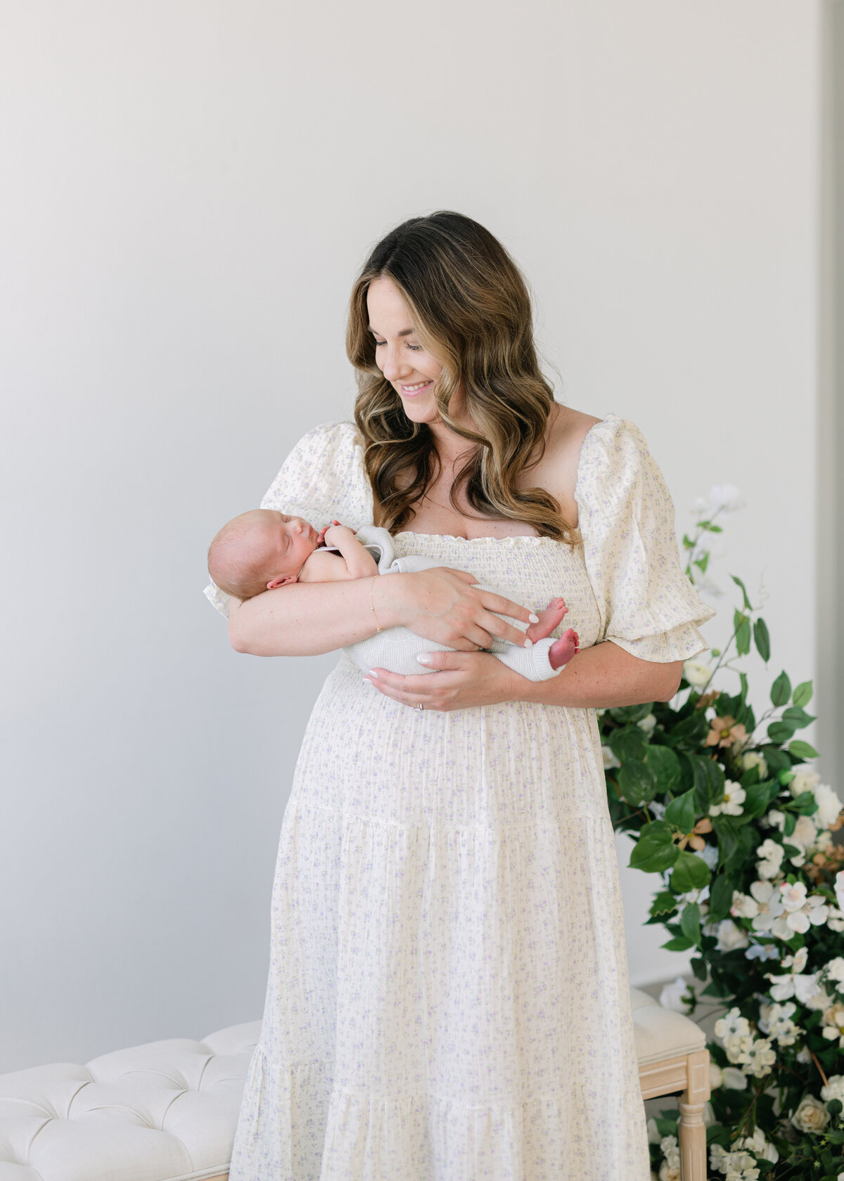 A mother in a long cream dress holing her newborn baby in front of a floral arch and tufted bench