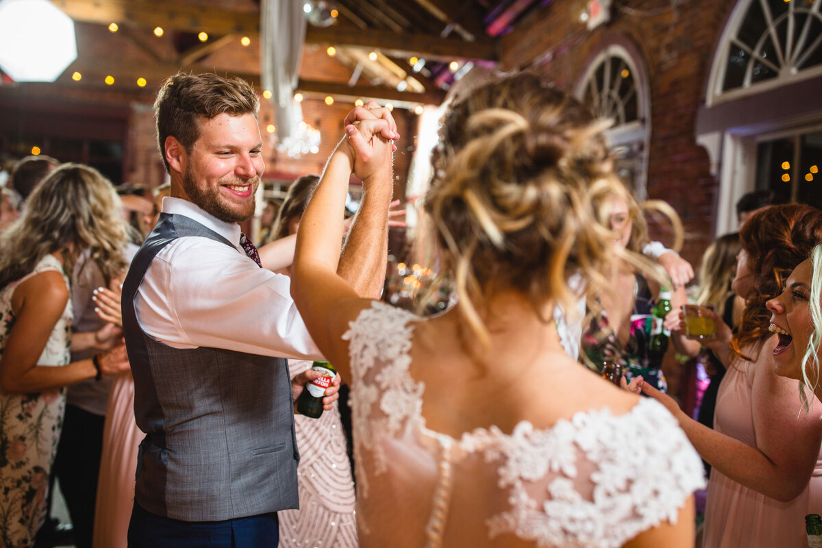 A joyful bride and groom share a dance at their wedding reception in Columbus, captured by a skilled Columbus wedding photographer. The groom, in a stylish grey vest and tie, smiles broadly as he twirls his bride, whose elegant lace wedding dress swirls around her. Her hair is styled in a loose updo, accentuating the delicate lace details on her dress. The warmly lit venue, adorned with string lights and a rustic brick backdrop, buzzes with the excitement of friends and family celebrating around them.