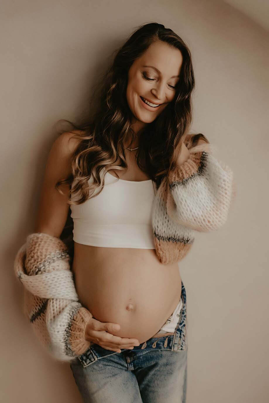A pregnant mother holds her belly while touching her hair and smiling