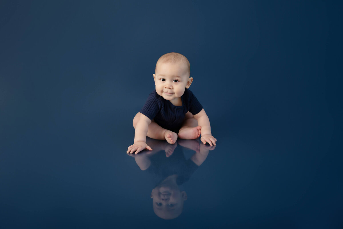 Baby boy wearing a navy blue onesie on a blue background crawling towards the camera for his Burlington, Ontario kids family portrait session