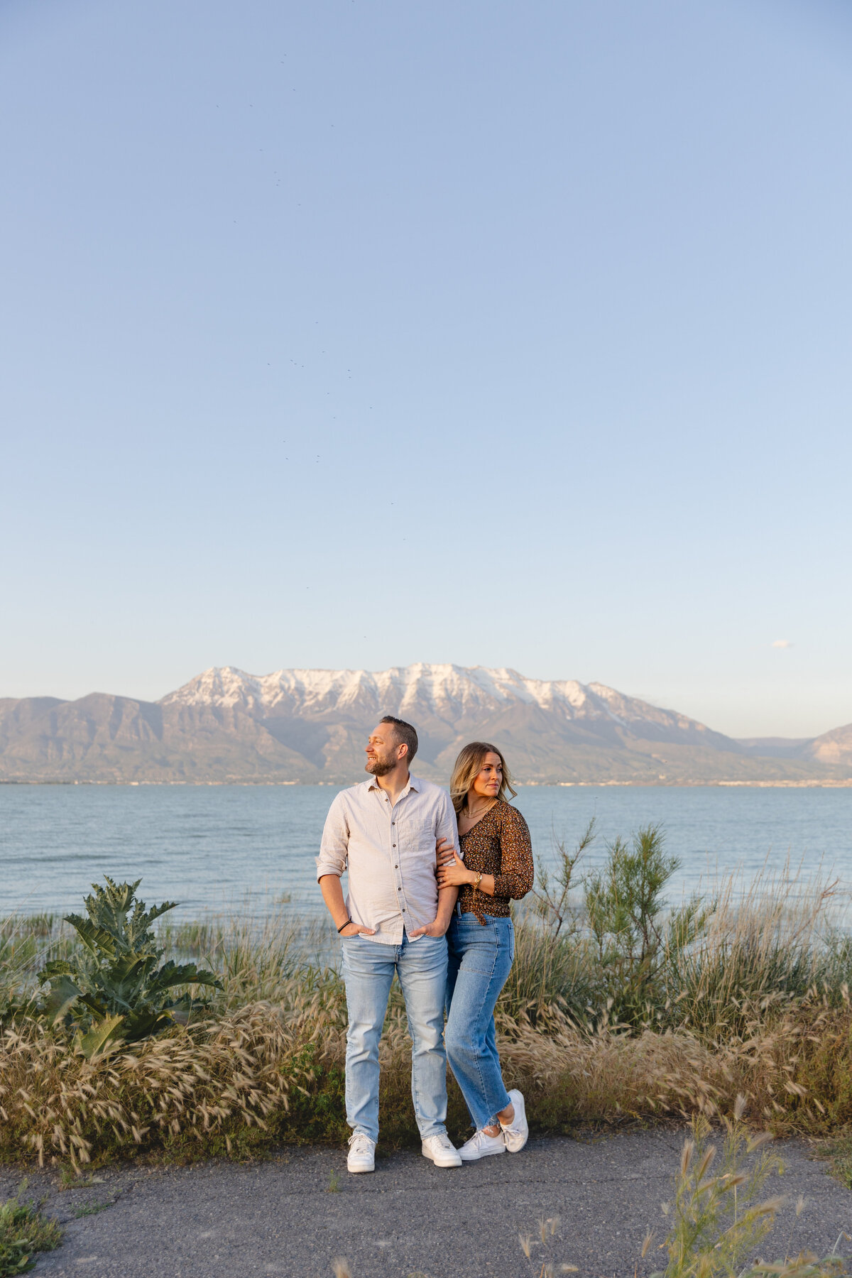 A couple poses by Utah Lake with the Wasatch Mountains in the background. The woman rests her hand on her fiancé’s arm as they look off into the distance, bathed in golden hour light.