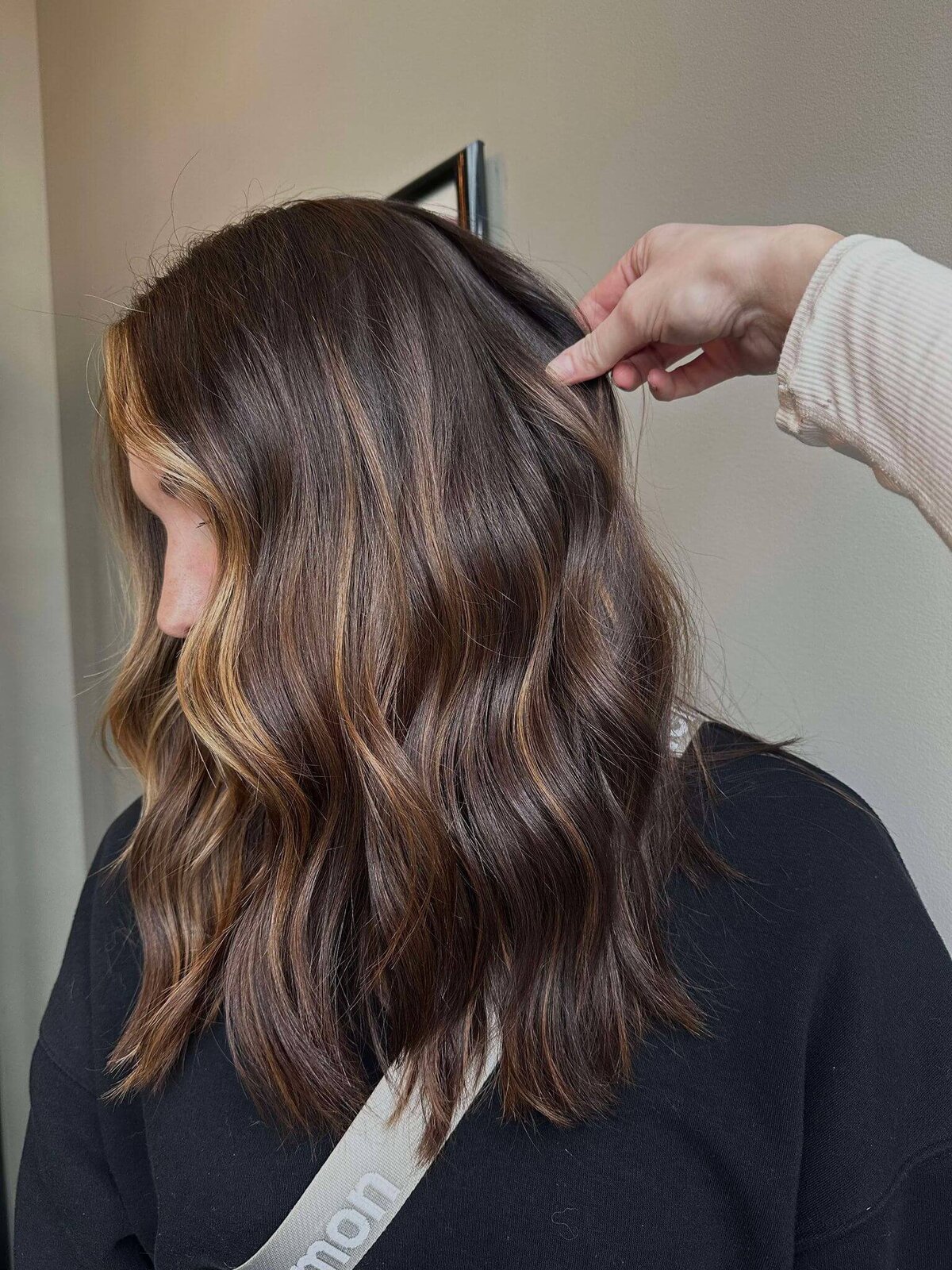 A woman with long, wavy brunette hair featuring subtle highlights, with a hand gently adjusting her hair, showcasing a luxurious style created by 212 Salon, Spa, & Barbershop.