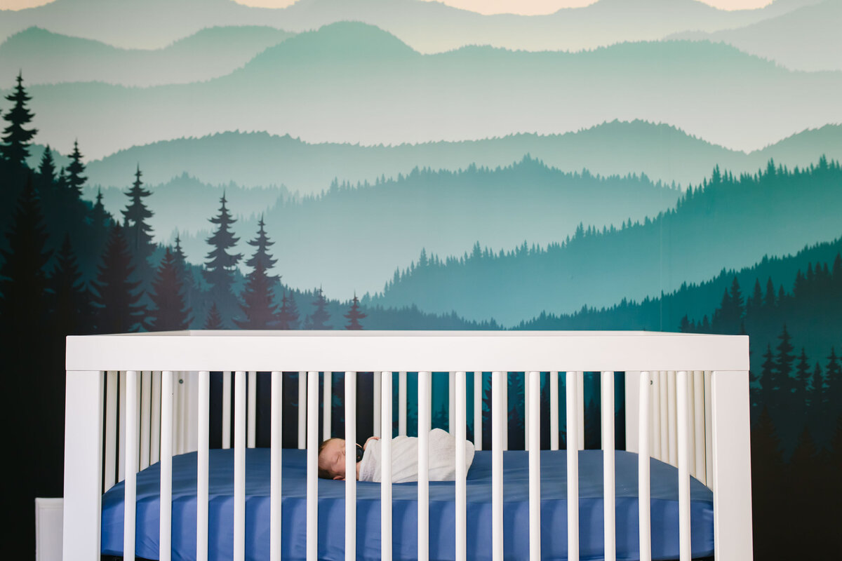 newborn baby laying on back swaddled in crib with mountain mural on nursery wall