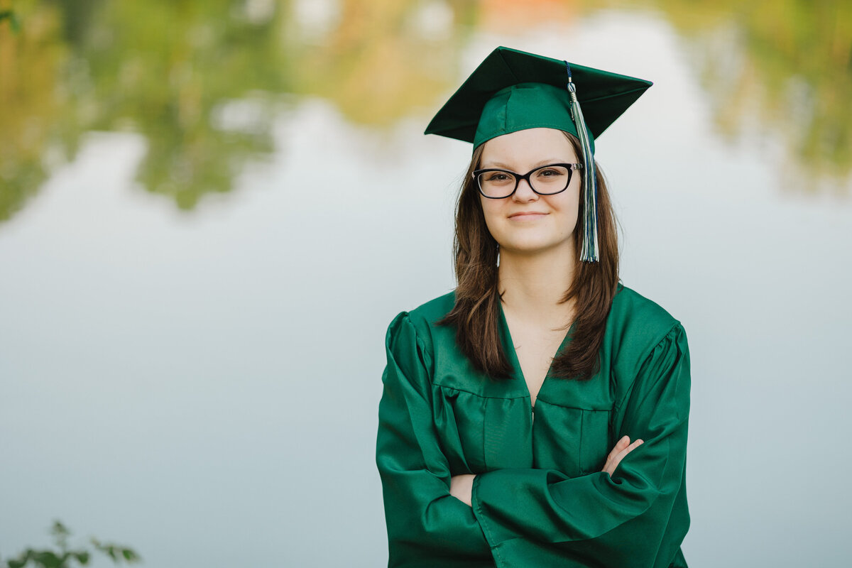 A graduate poses with arms crossed in a green graduate outfit outdoors.