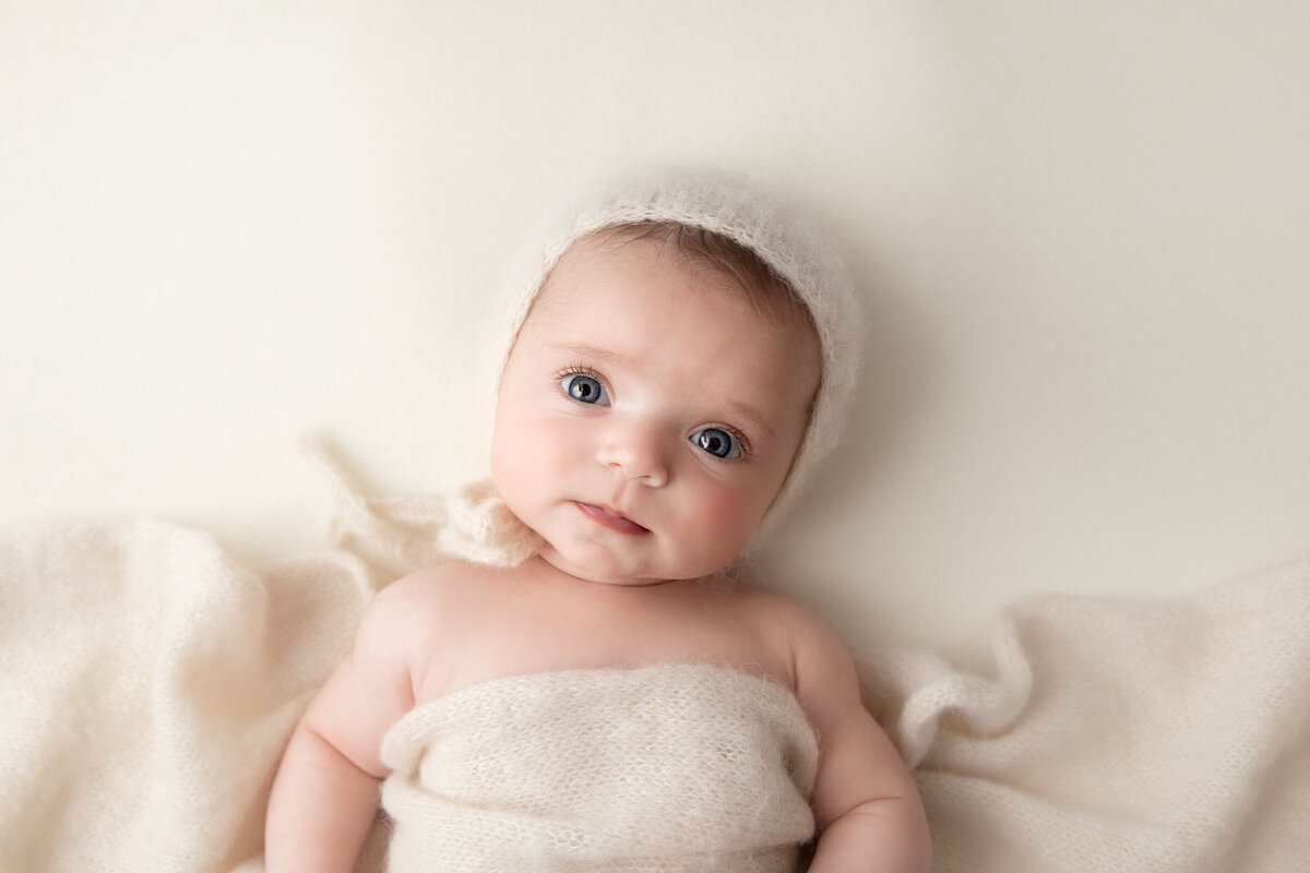 Baby girl looking sweetly at the camera wearing a cream knitted bonnet and a matching knitted wrap laid over her.