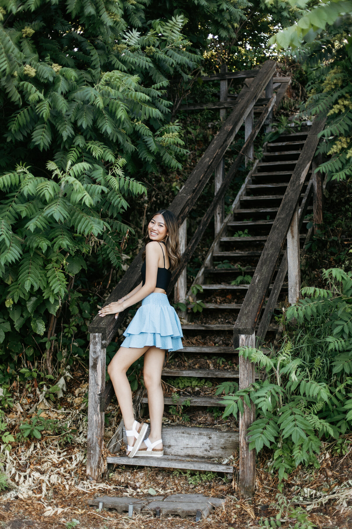 Senior girl in a black top and blue skirt posing on a rustic wooden staircase surrounded by greenery