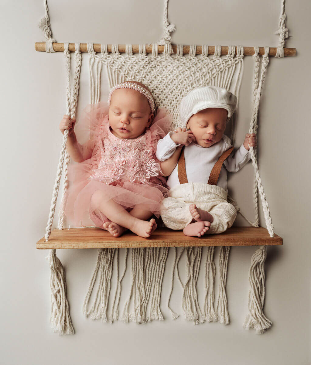 Baby twins newborn age, boy  and girl sitting on a macrame swing dressed in a pink dress and white overalls and hat.