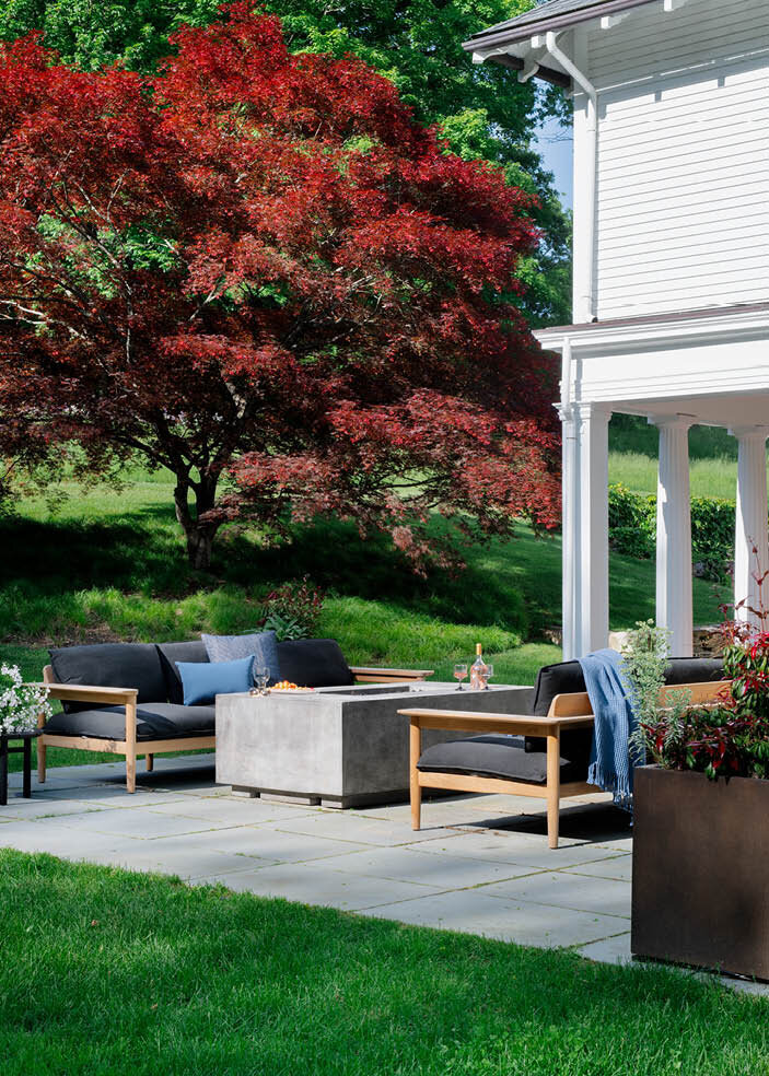 View of the bluestone patio featuring a gas firepit, teak sofas from Design Within Reach, and small Ethnicraft side tables. As seen on Boston Home Magazine's Instagram.