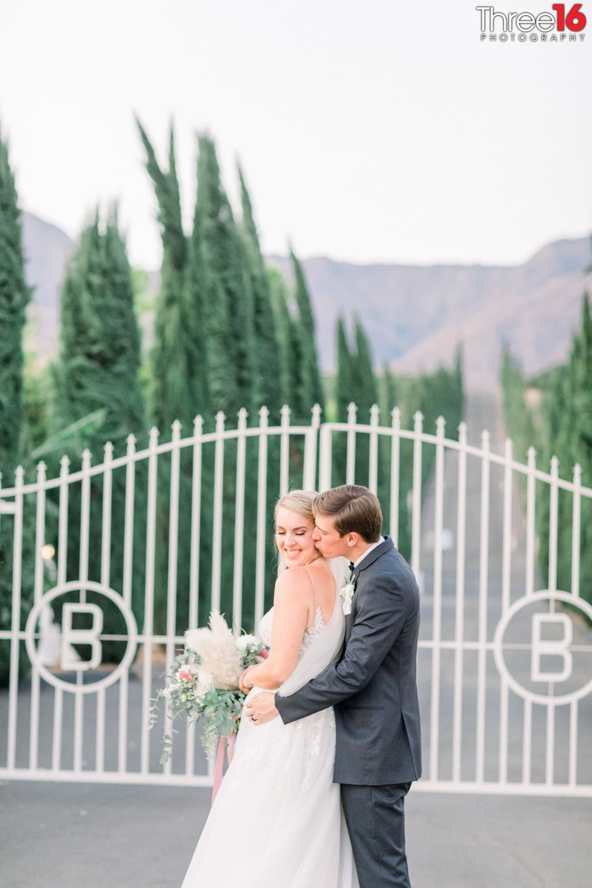 Groom embraces his Bride from behind and kisses her cheek leaving her a big smile