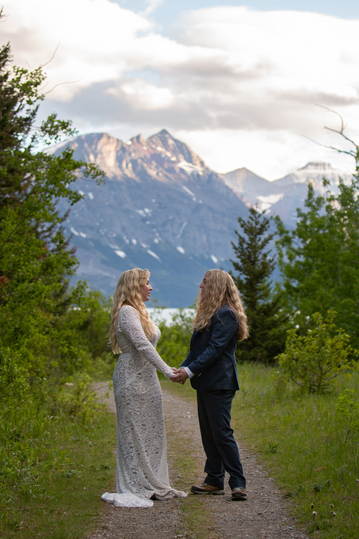 Two brides stand holding hands and facing each other on a dirt path in Glacier National park as the sunset in the mountains behind them.