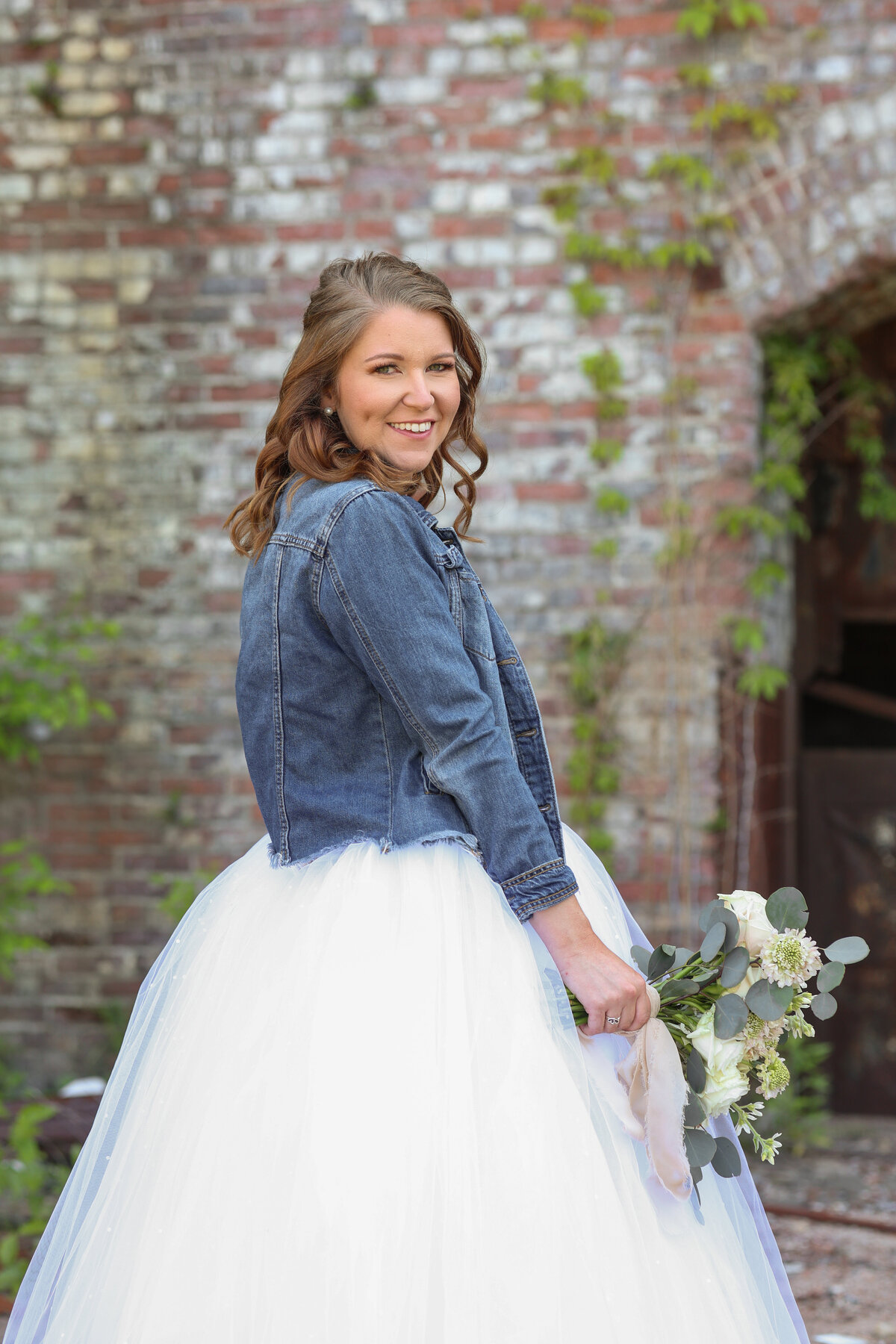 bride wearing blue jean jacket and holding white floral bouquet at The Bibb Mill by Columbus Georgia wedding photographer Amanda Richardson Photography