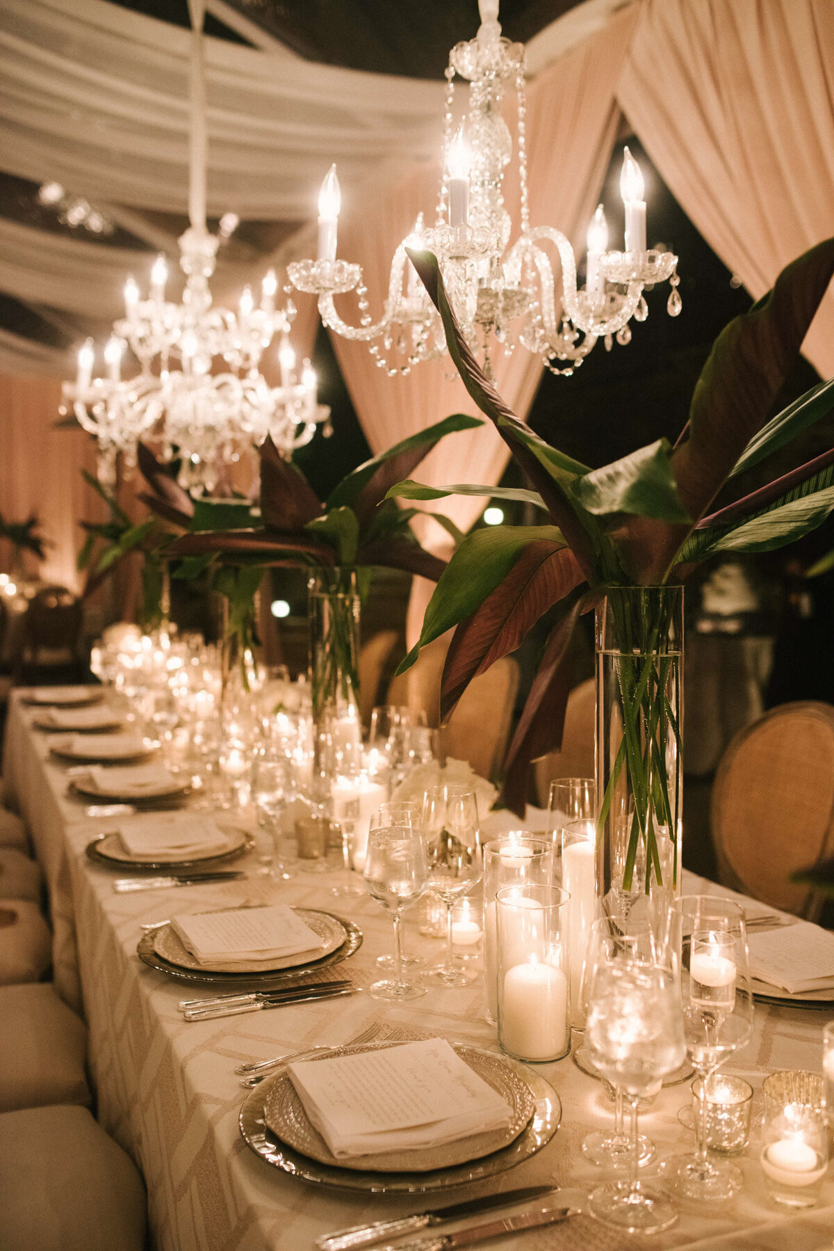 Elegant wedding reception rectangular table with botanical centerpiece, lighted candles in a glass, plates, utensils and glasses