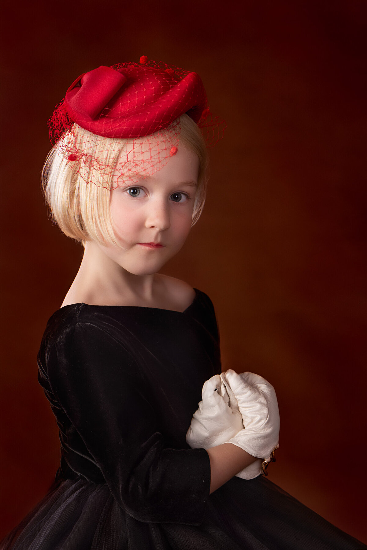 Sophisticated fine art portrait of a young girl wearing a vintage red hat and white gloves, posed against a deep red backdrop. Captured in Overland Park, Kansas, this timeless setup is ideal for families seeking refined and artistic child photography.