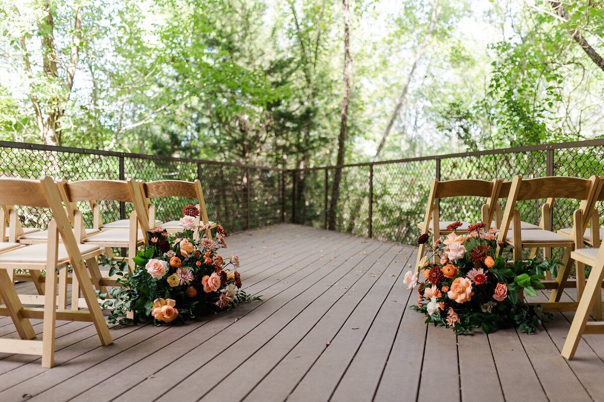 A detail shot of the ceremony space for a wedding at the Trinity River Audubon Center in Dallas, Texas. The front rows of chairs be seen from the aisle point of view and the second row of chairs have large bouquets on flowers on either end. The ceremony space overlooks lots of greenery and nature.