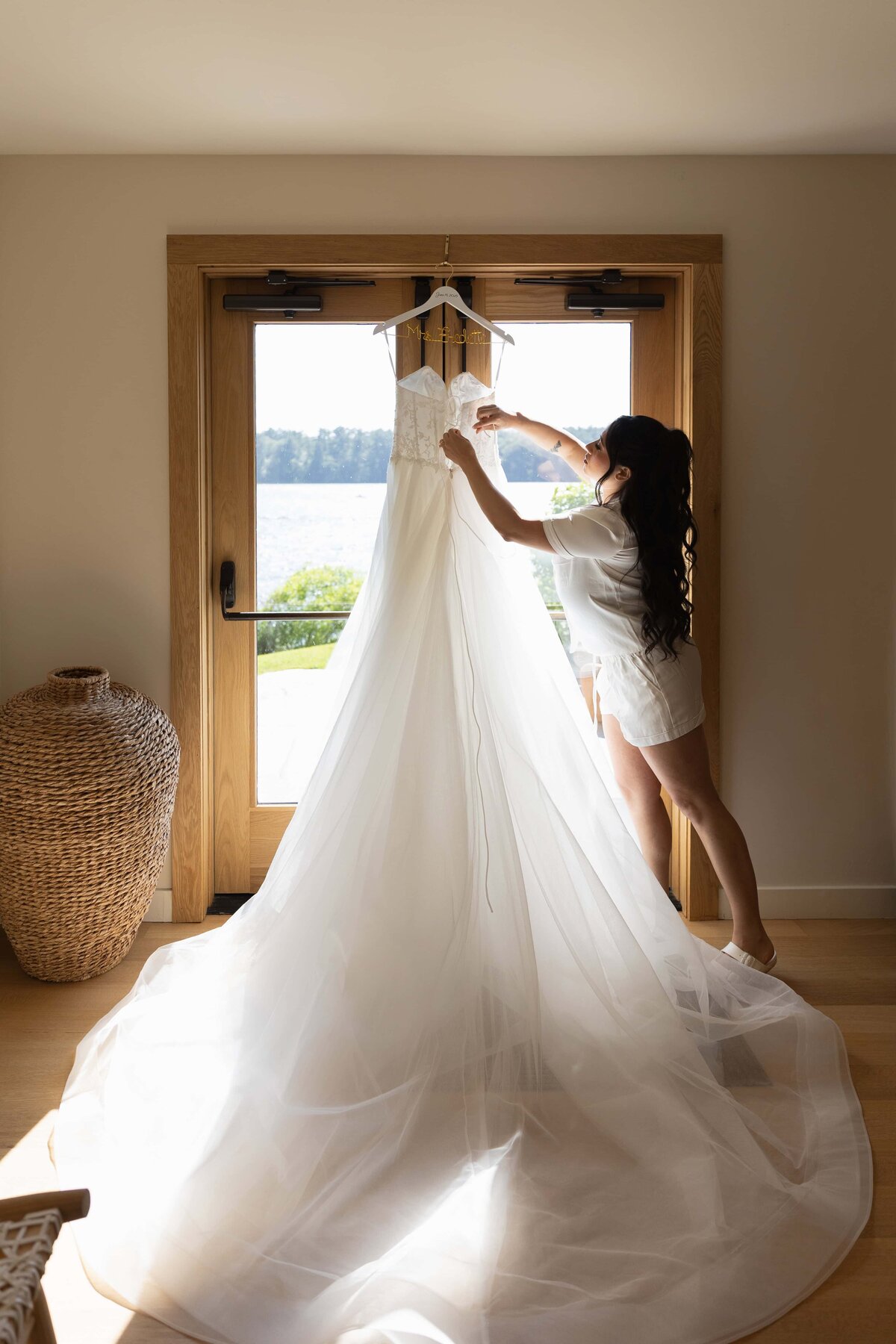 The Lakehouse Inn MA  | Kelsey Sheehan Photography Timeless Rhode Island Weddings | A woman in white pajamas admires a flowing wedding dress hanging on a door. Sunlight streams in, highlighting a serene lake view outside, creating a calm and joyful atmosphere.