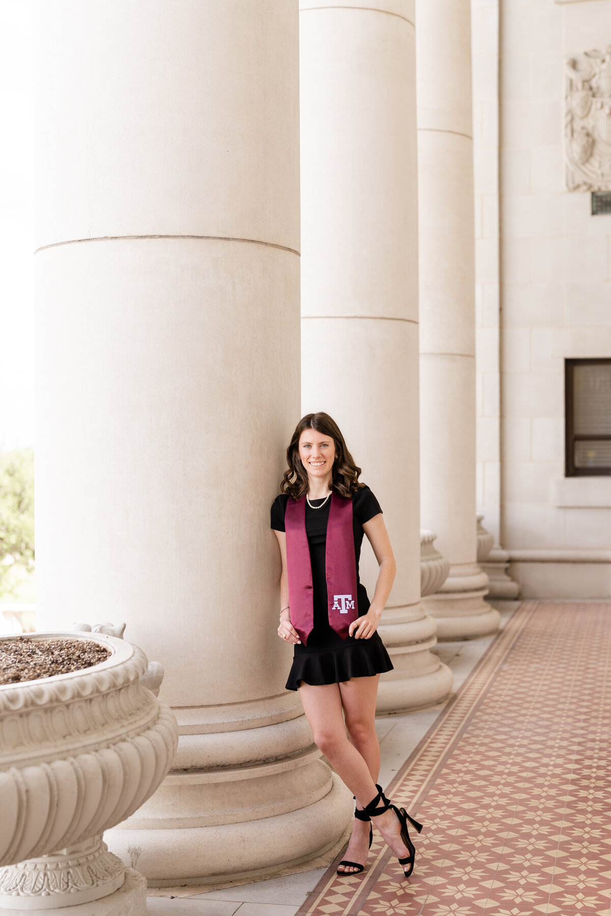 Texas A&M senior girl holding stole and leaning against column while wearing black dress and maroon stole at Administration Building