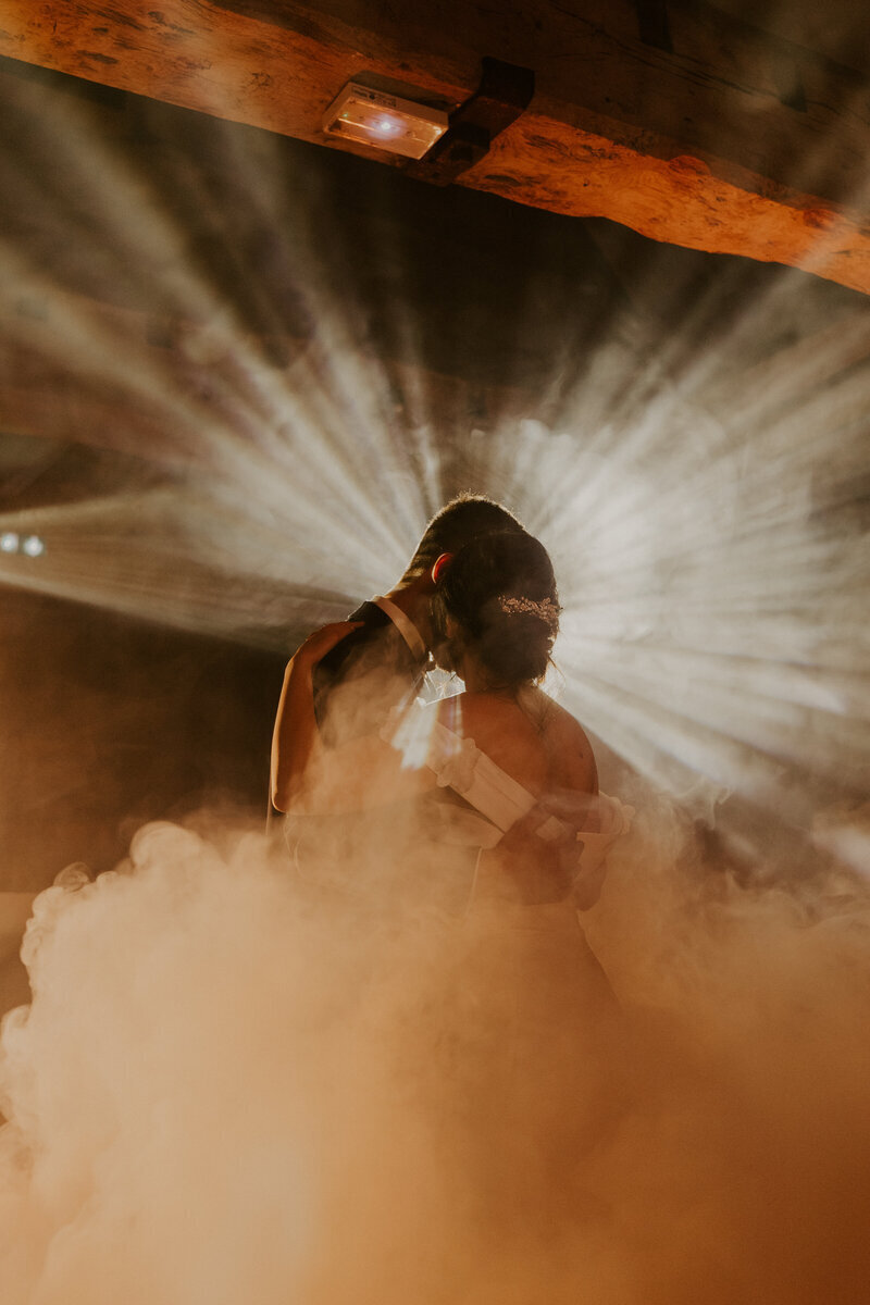 Couple de dos pendant l'ouverture de bal, sous les projecteurs, ambiance tamisée et fumée beige orangée. Photo prise pour le shooting photo mariage.