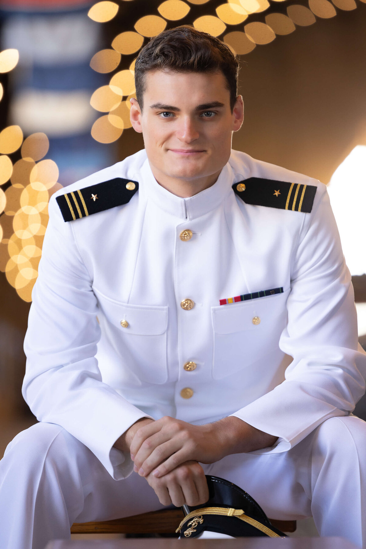 Naval Academy Midshipman in choker white uniform sitting on a bench and smiling with golden lights behind him.