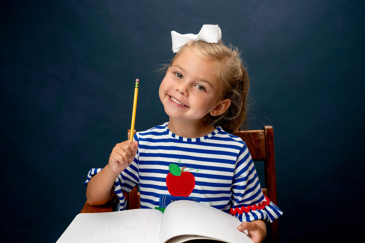 Young girl in blue and white striped sitting at desk with journal and pencil smiling.