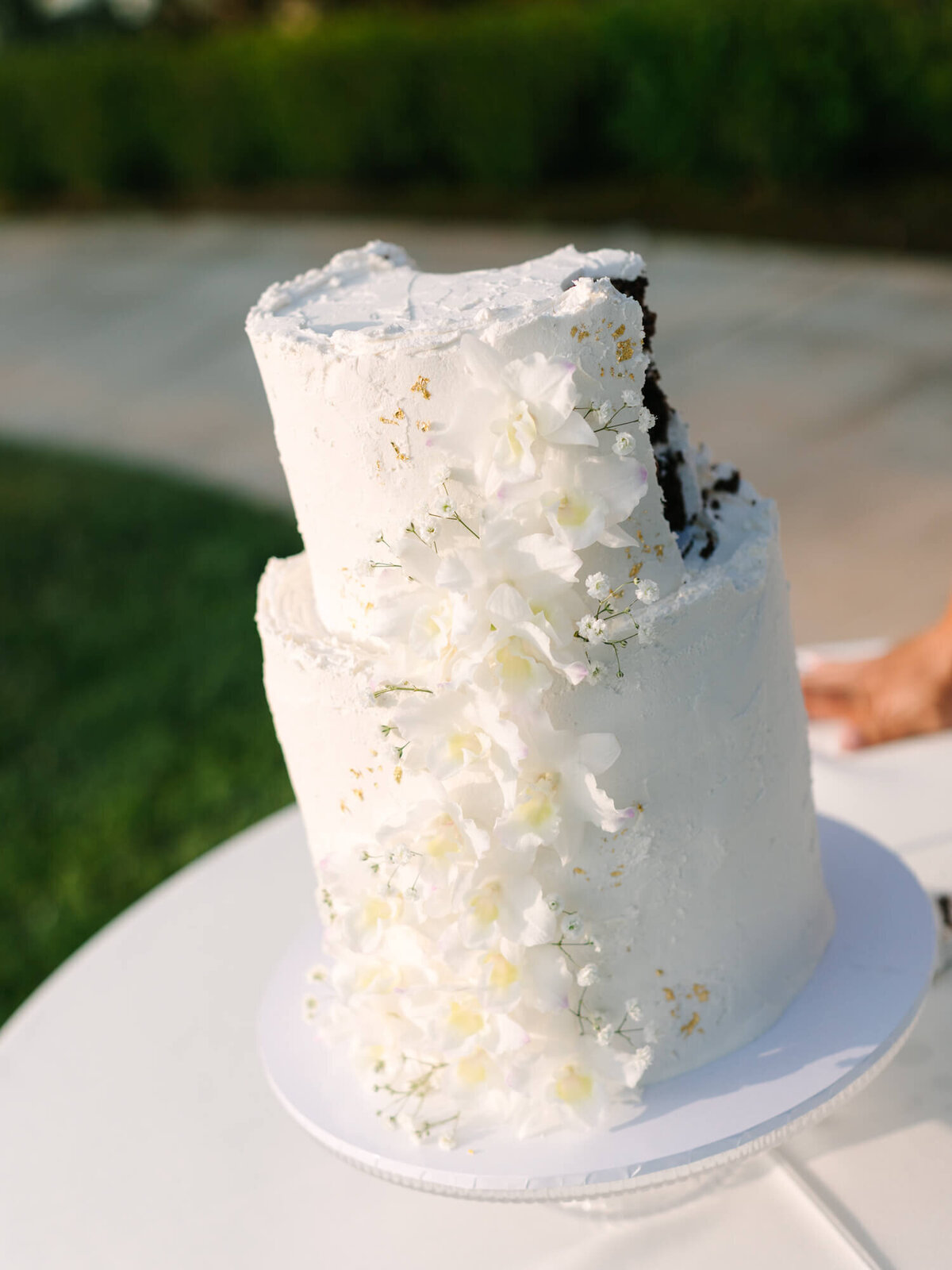 A two-tiered white frosted cake with cascading white flowers and has a slice cut out, revealing dark chocolate inside.