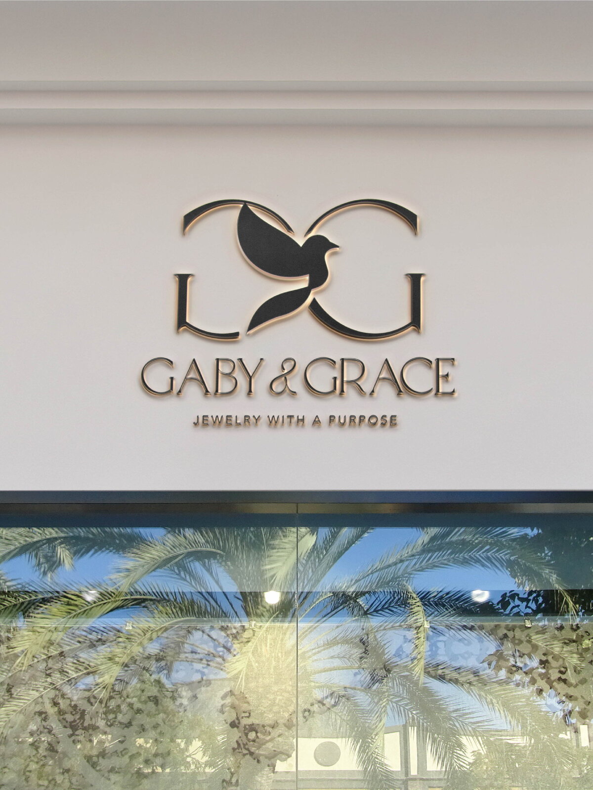 Elegant storefront signage mockup for Gaby & Grace featuring the monogram dove icon and brand name in dimensional lettering, with the tagline “Jewelry With a Purpose” mounted on a clean off-white facade above glass doors reflecting palm trees.