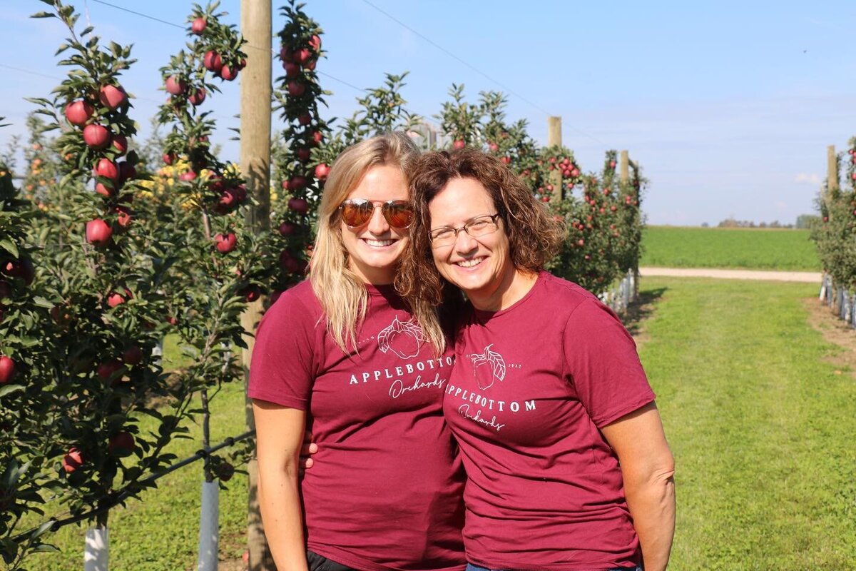 Chelsea, owner, and her proud mother smiling while sporting Applebottom Orchards shirts in the orchard.