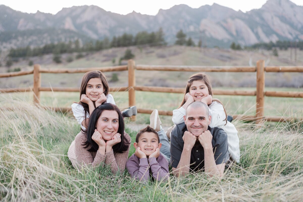 A mom and dad lay on the ground on their stomachs with their daughters each on their backs with their hands tucked under their chins and the son in the middle between everyone on his stomach too