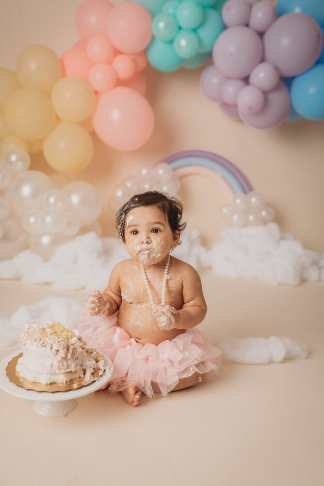 Photo shoot at Atlanta, Georgia cake smash photographer studio with one year old little girl in pink tutu smashing a cake with rainbow pastel balloon garland behind her with white fluffy clouds.