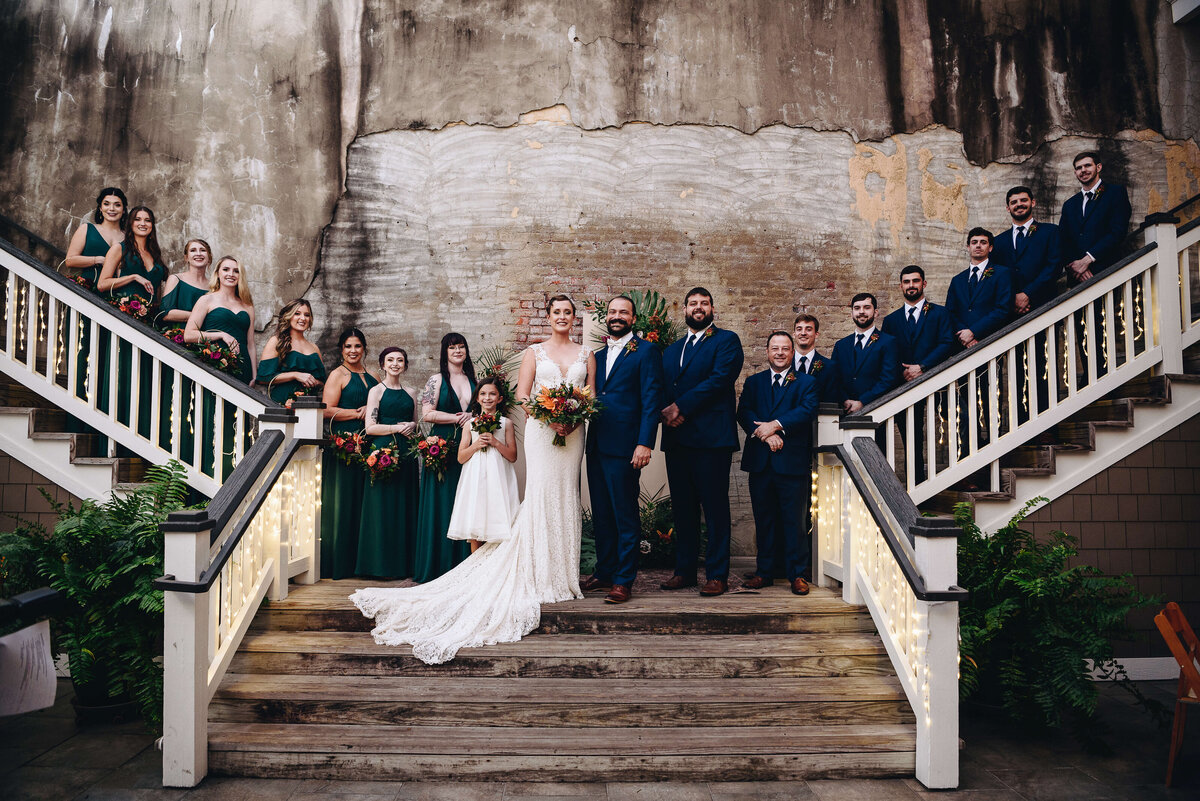 A wedding party poses on a staircase in front of a brick wall. The bride and groom stand at the center, surrounded by bridesmaids in green dresses and groomsmen in navy suits, all holding bouquets or standing with hands clasped.