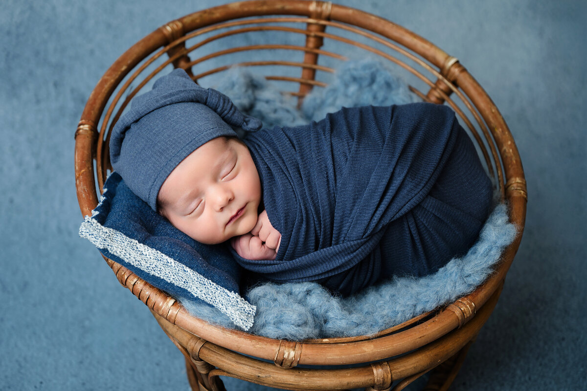 This sweet newborn photography session in Pittsburgh features a baby boy peacefully swaddled in a soft blue wrap, resting in a wicker basket. The neutral tones and soft textures highlight the serene beauty of a newborn’s first portrait.