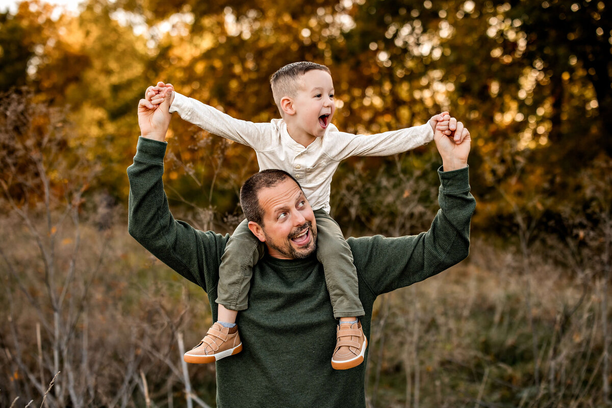 A man in a green sweater carries a joyful child on his shoulders. The child is wearing a light shirt and green pants, holding the man's hands. They are outdoors with blurred autumn foliage in the background.