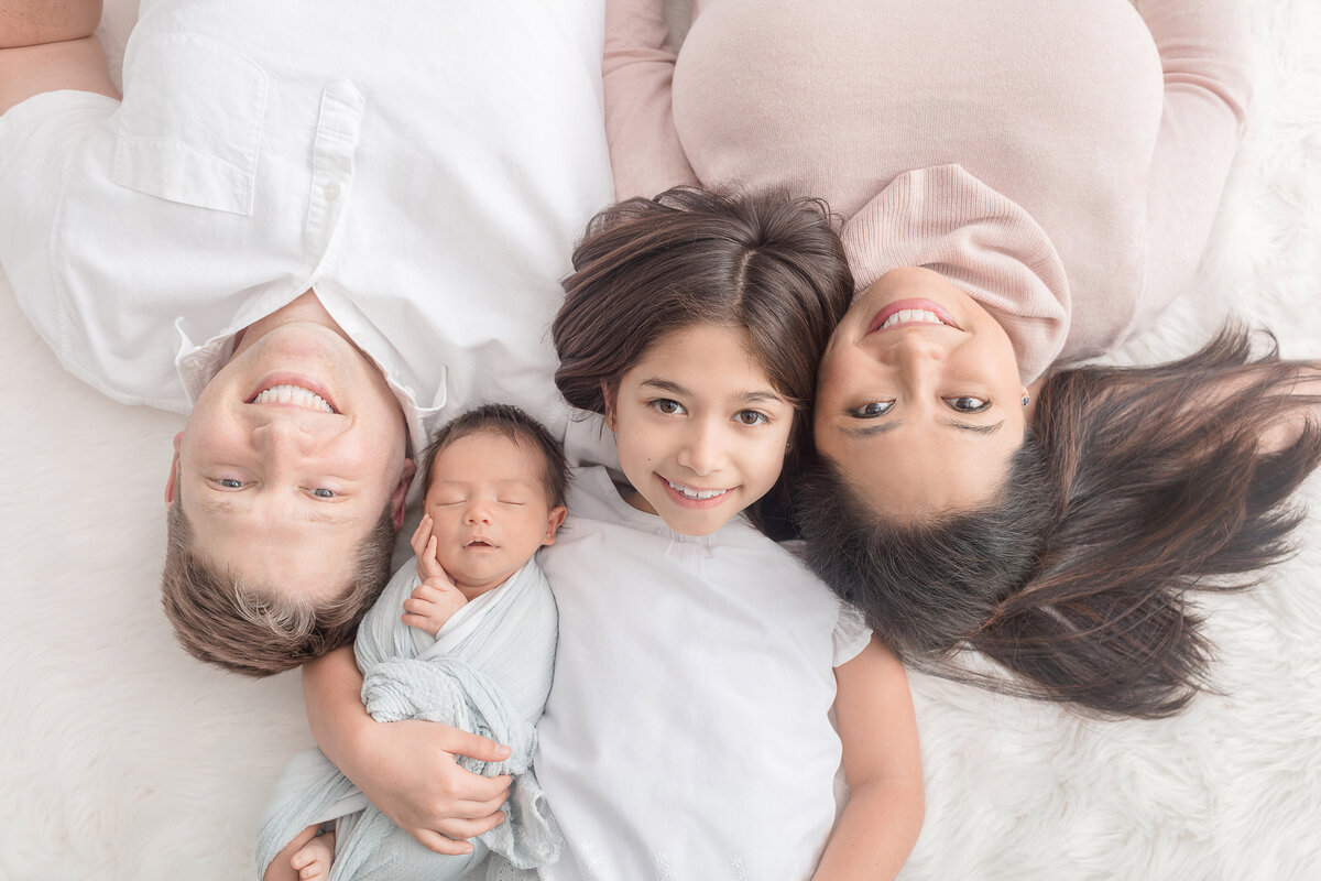 A new family of four lie on a white blanket in the Looking Up Photography photo studio. The family are smiling, with the exception of the newborn who sleeps peacefully, touching their face with their hand, with a slight smile on their face.