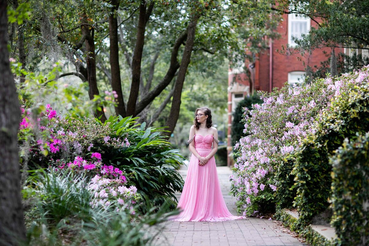 senior in pink gown on sidewalk surrounded by flowers