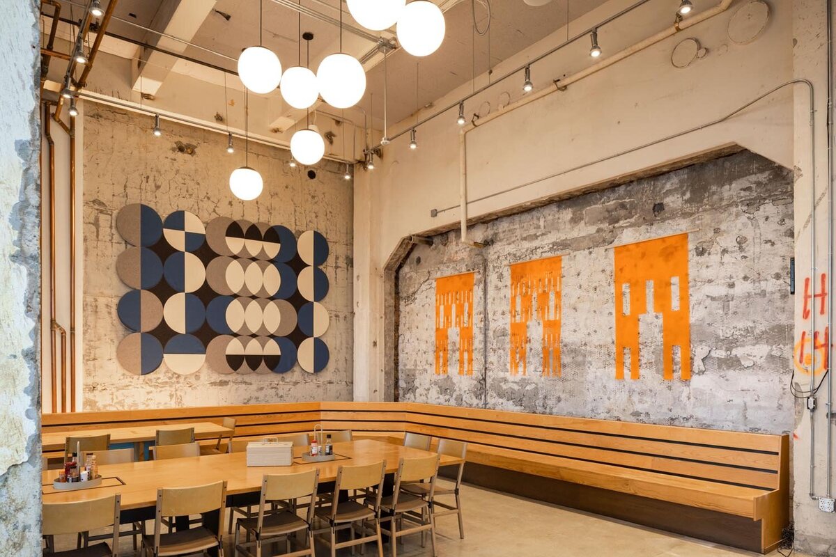 Industrial-style dining and gathering area with a long wooden table, wood bench seating, exposed concrete walls, geometric wall art, and globe pendant lighting.