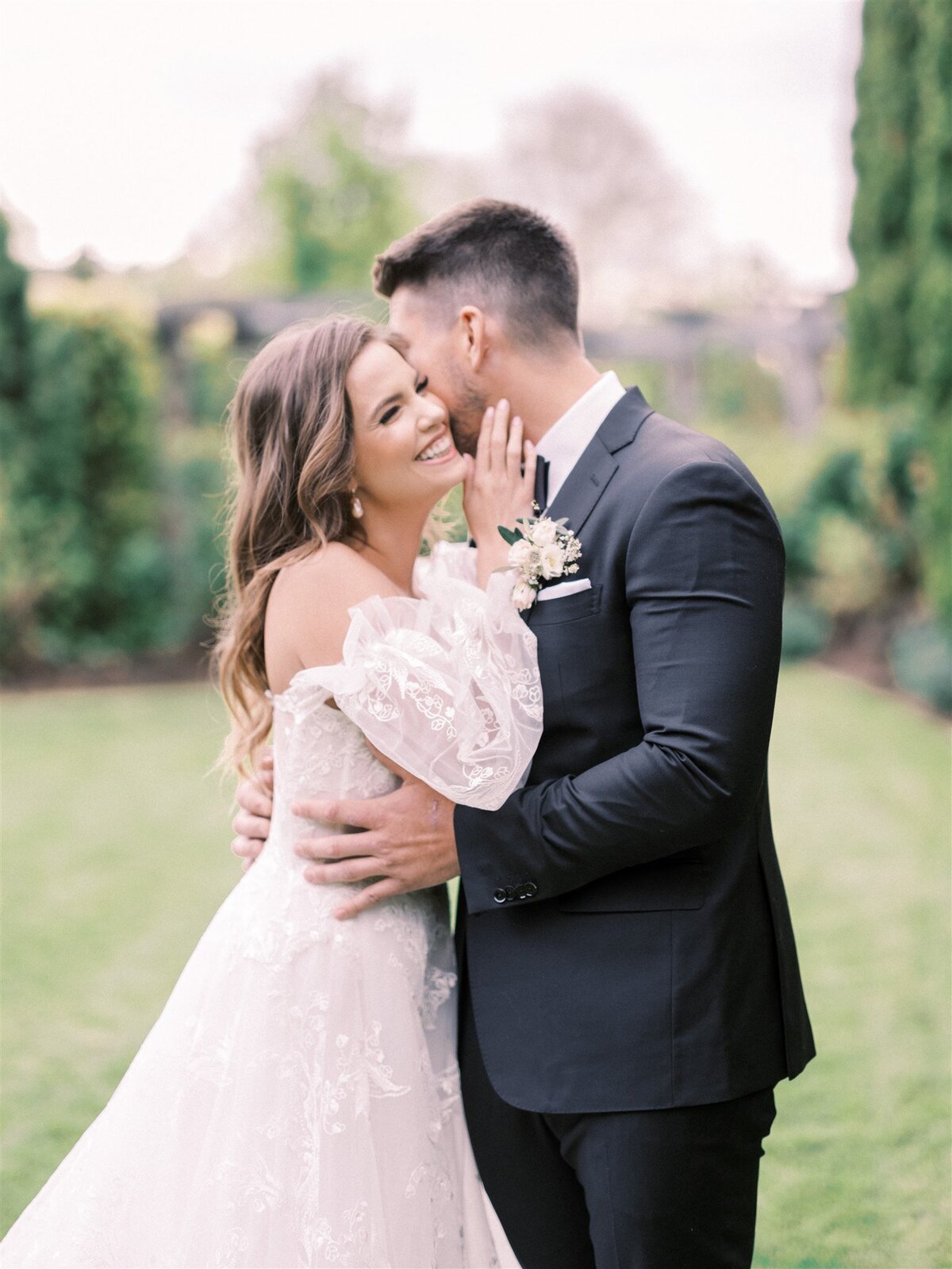 A bride and groom embrace in an outdoor setting, arranged by Destination Wedding Planner Melissa Dawn Event Designs. The bride, in an off-the-shoulder lace gown, smiles while the groom, in a dark suit, gently holds her face. Trees and greenery are in the background.