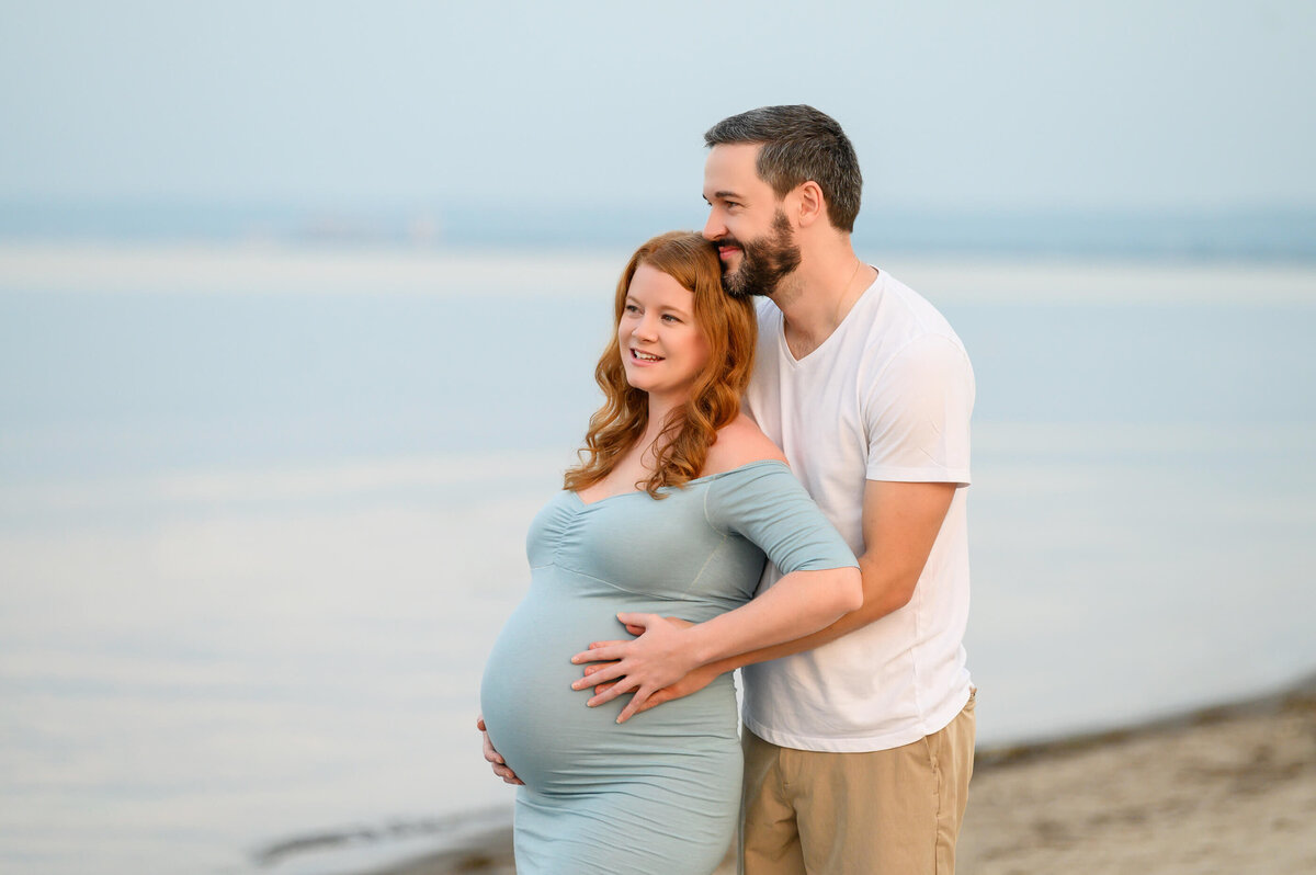 mom and dad holding pregnant wife's belly looking out into the water for their beach maternity photography session in Burlington, ON 