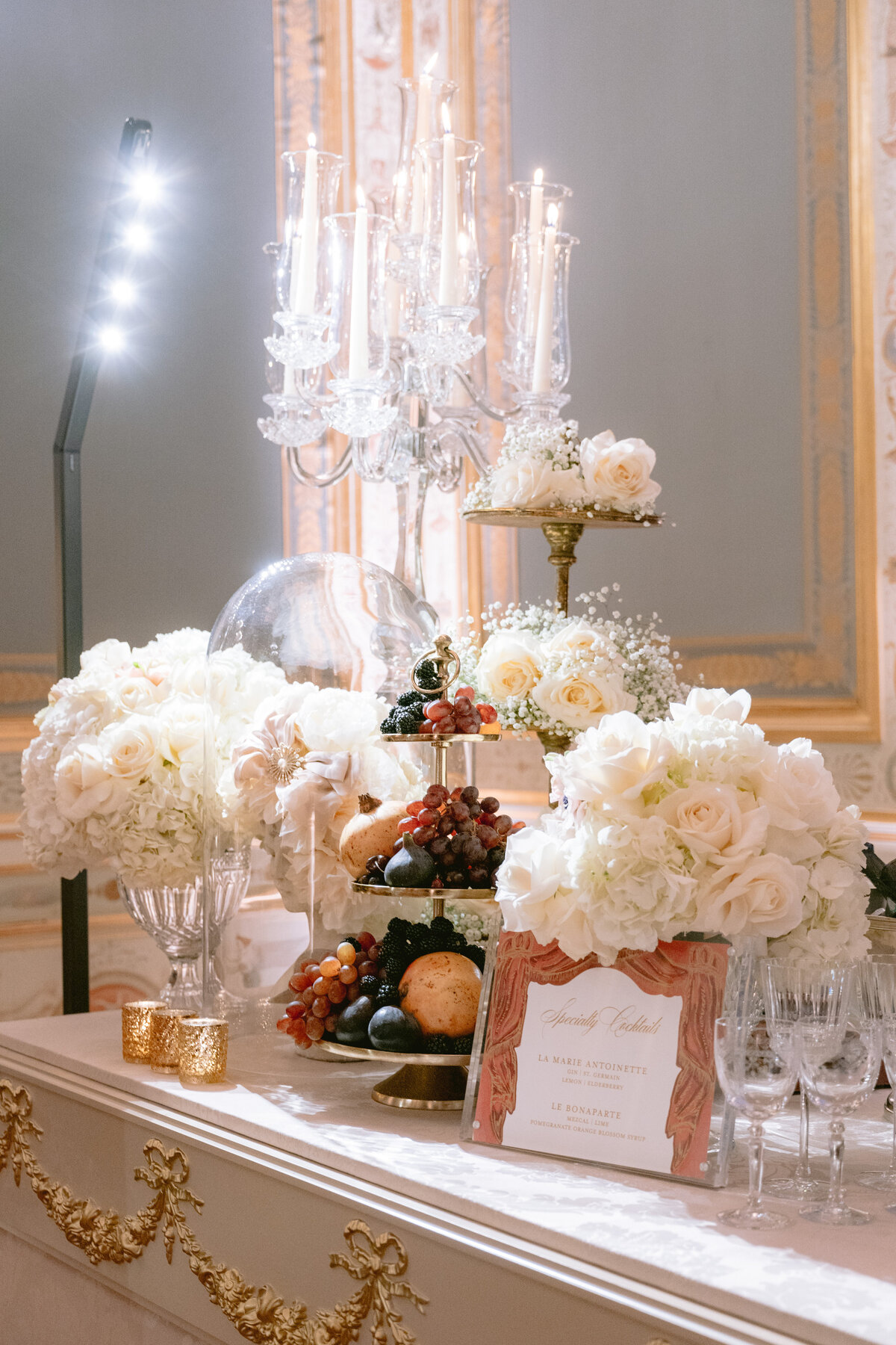 The photo was taken at Shangri La Hotel & Ritz Paris Hotel in Paris, France by photographer Matteo Coltro, with videographers Zen Film Works capturing the scene. The image showcases a lavish table setup featuring a tiered stand adorned with luscious grapes, figs, and assorted breads. Alongside, elegant white roses are delicately arranged, crystal glasses glisten in the soft lighting, a decorative candelabrum adds a touch of sophistication, and an ornate card completes the opulent display. The gold accents further elevate the luxurious atmosphere, creating a truly exquisite setting.
