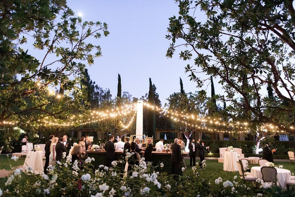 Outdoor wedding reception space at dusk, there are twinkling lights over the space, and guests are walking arounf.