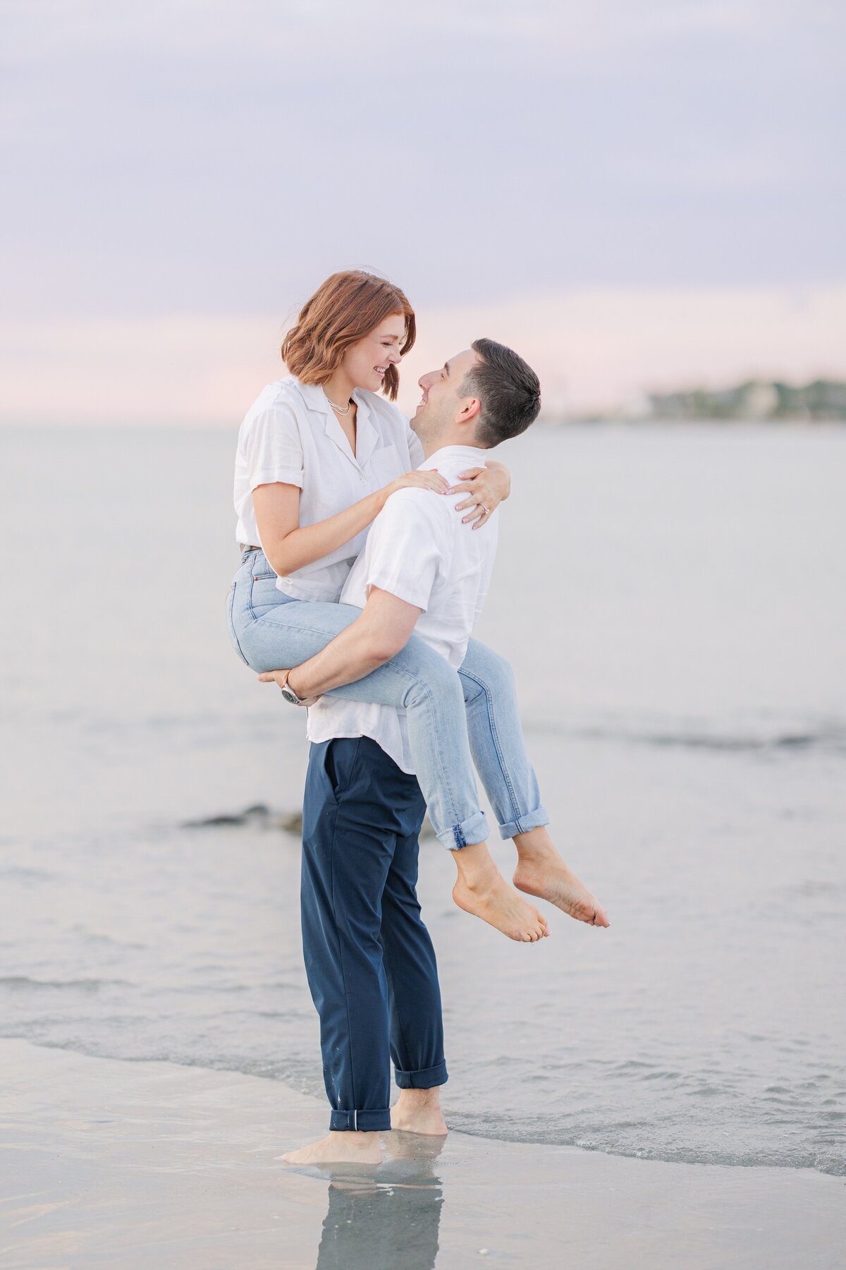 A man picking up and snuggling a woman during their Wingaersheek Beach engagement session.